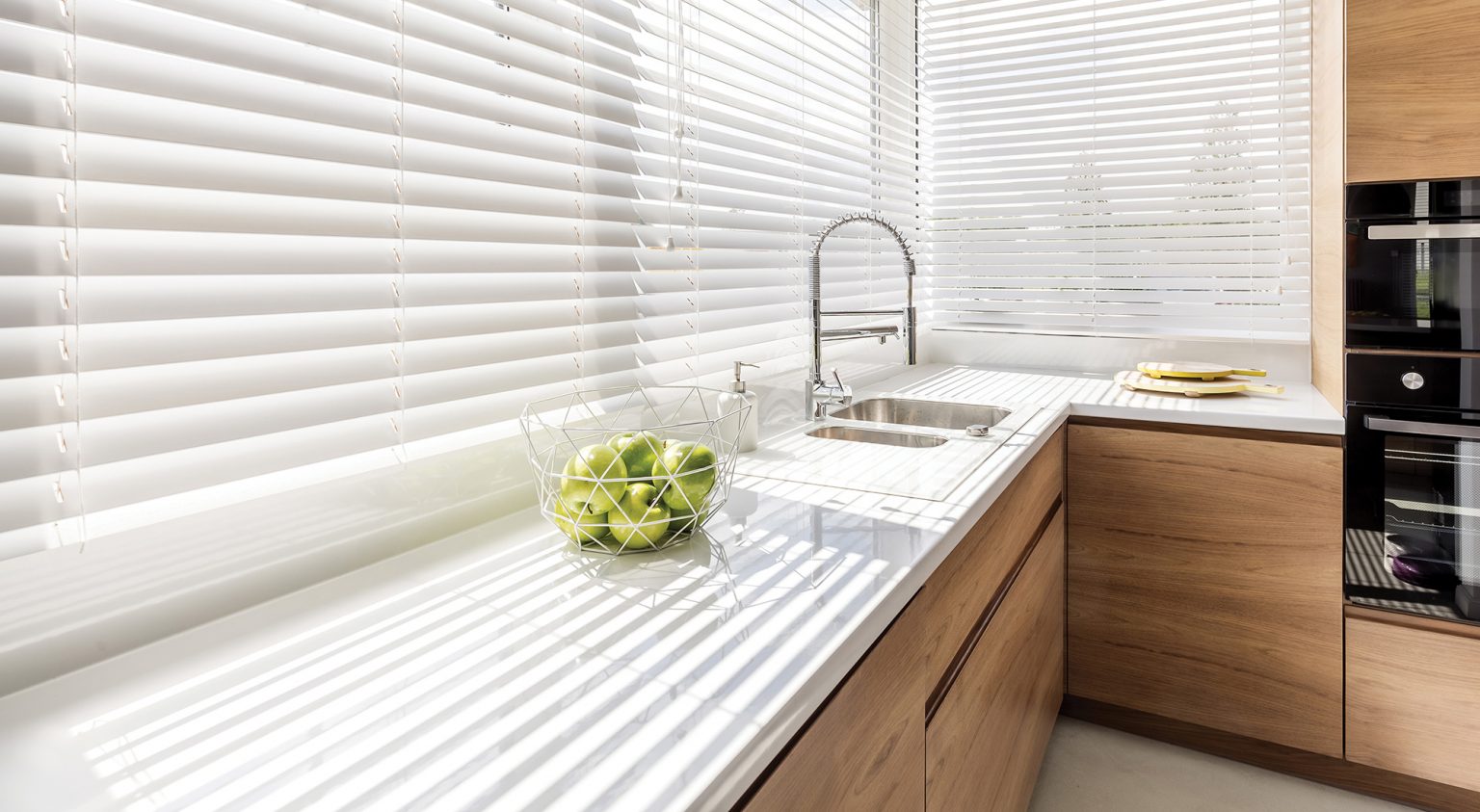 Modern bright kitchen interior with white horizontal window blinds, wooden cabinets with white countertop and household appliances